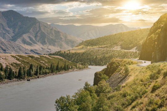 Wide landscape view of Chuysky Trakt road following the Katun River through mountain valley at golden hour, Altai Republic, Russia, with dramatic clouds and summer greenery