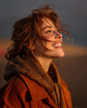 A young woman with red hair smiles joyfully as the wind tousles her hair, wearing warm autumn clothing against a blurred natural background at sunset.