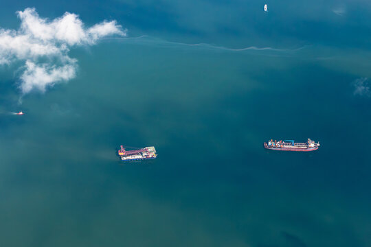 An aerial, top-down view captures a large dredging vessel and a tanker ship navigating the blue ocean. White clouds float above the calm waters, showcasing maritime industry activity.