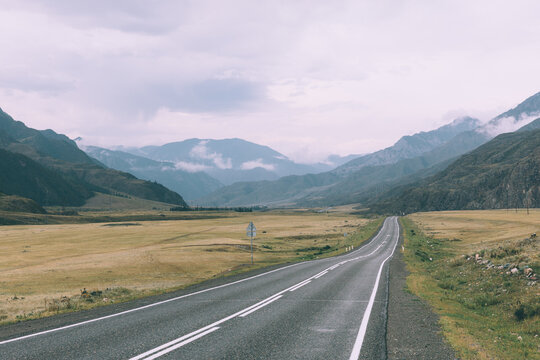 Empty asphalt road of the Chuysky Trakt stretching through a wide mountain valley with meadows and low clouds, Altai Republic, Russia, travel landscape background