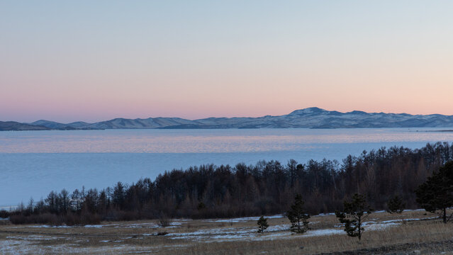 Wide winter landscape with frozen Lake Baikal in the Maloye More strait, pastel sunset sky, distant snowy mountains and forested shoreline, Siberia, Russia