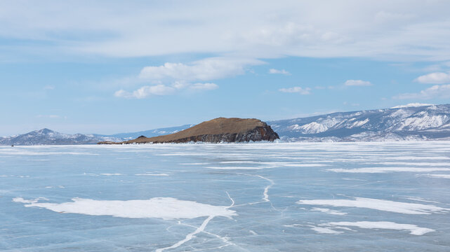 Wide winter view of frozen Lake Baikal with blue ice, snow patches and natural cracks, with a rocky island and mountains on the horizon, Russia, Siberia