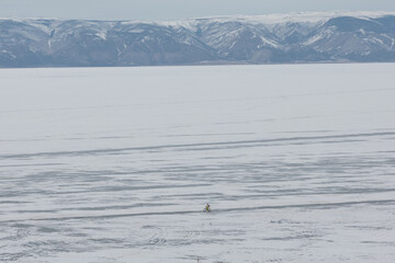 Naklejka premium Wide winter landscape of frozen Lake Baikal with a lone cyclist riding across the ice, distant snow-covered mountains and overcast sky, Russia, Siberia