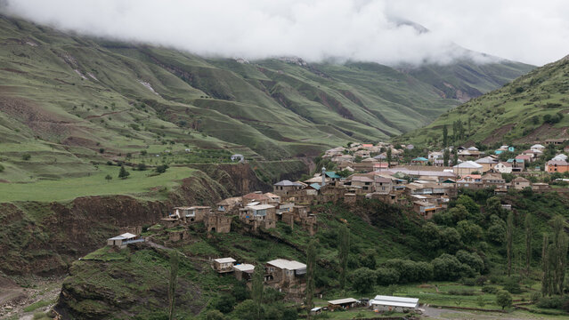 Panoramic view of Magar village on a hillside in Dagestan, Russia, with traditional houses, lush green mountain slopes, and misty clouds over the valley