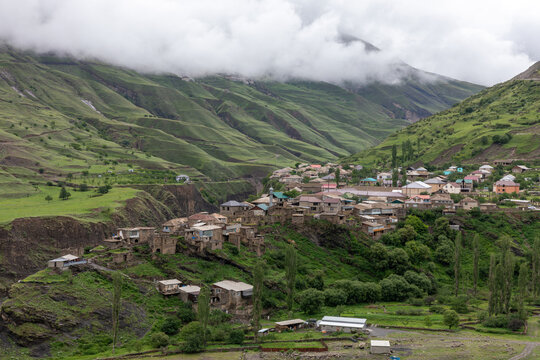 Wide view of Magar village in Dagestan, Russia, with traditional houses on a hillside, lush green mountain slopes, winding roads and dramatic low clouds