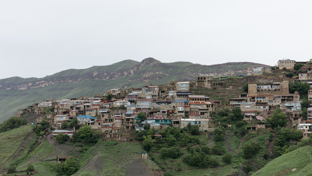 Wide panoramic landscape of Chokh aul with traditional houses stacked on a steep green slope, mountain ridges and cloudy sky in Dagestan, Russia