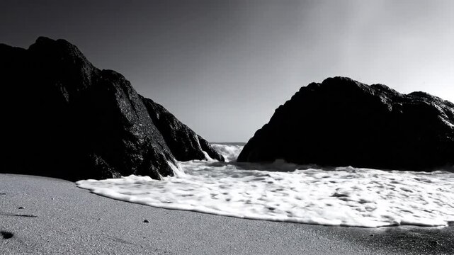 Dramatic black and white ocean waves crashing between rugged rock formations on a sandy beach under an overcast sky.