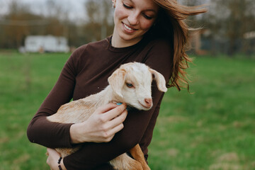 Fototapeta premium Smiling woman hugs a goat kid in a grassy field, highlighting a gentle countryside lifestyle, connection with animals, and pastoral calm under soft natural light, creating a warm, hopeful mood.
