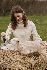Fototapeta premium Joyful woman outdoors feeding a goat kid on a hay bale, sunlit field, cozy knit sweater, gentle interaction, soft breeze, natural colors, rural lifestyle moment