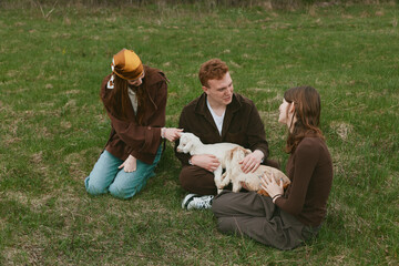 Fototapeta premium Goat, kid, friends, outdoor, park, petting, animal, family describe a candid lifestyle scene as three people softly interact with a gentle goat kid on a grassy field, smiles, relaxed clothing,