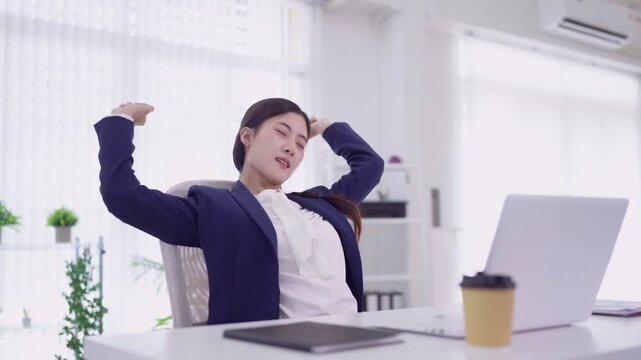 Asian businesswoman stretching arms relaxing at desk during work break using laptop computer in bright modern office taking rest from corporate professional