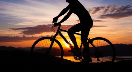 Obraz premium Silhouette of a person cycling a bicycle on a hill against a vibrant sunset sky over mountains and water.