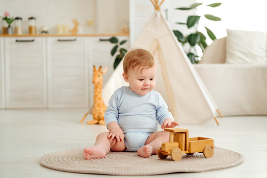 A smiling and joyful kid is playing on the floor with wooden toy cars near the wigwam in the children's room at home, a small child is sitting in a blue bodysuit laughing