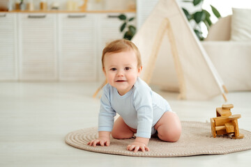 A smiling and joyful kid is crawling on the floor near the wigwam in the children's room at home, a small child is learning to crawl in a blue bodysuit and laughing © Any Grant