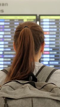 Backpack Traveler Facing Departures Board, Back View Woman With Ponytail And Gray Backpack Scanning Digital Flight Display In Busy Airport Concourse, Soft Neutral Lighting, Quiet Anticipation,