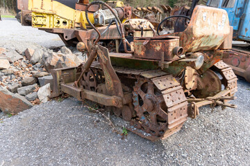 Fototapeta premium Old rusted crawler tractor with front blade, metal tracks, hydraulic hoses, engine parts, and worn seat near quarry rocks. The scene shows abandoned construction equipment, heavy machinery, industrial
