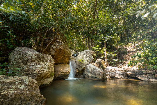 A peaceful natural landscape featuring a small waterfall flowing over large mossy boulders into a calm, clear rock pool, surrounded by dense green forest foliage.

