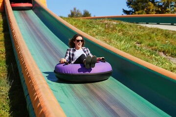 Obraz premium Woman smiles while sliding down tubing track on sunny day. She rides inflatable ring down green slope with grassy sides. Woman wears sunglasses and casual clothes, enjoying fun activity at resort.