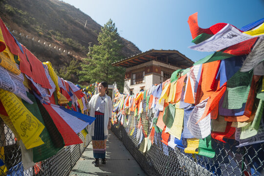 Tourist or Traditional Bhutanese suspension bridge adorned with vibrant prayer flags crossing a river, with a classic white building and rugged mountains in the background