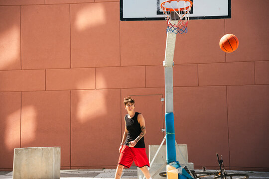 A young person in a black tank top and red shorts is on an outdoor basketball court. A basketball floats near the hoop. A bicycle rests on the ground. They look at the camera.