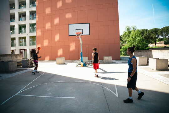 Three young adults enjoy a sunny day playing basketball on an outdoor court. One person prepares to shoot, another waits near the hoop, and a third walks across the concrete surface.