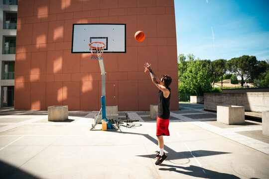 A young man jumps to shoot a basketball on an outdoor court during a sunny day. He wears red shorts and a black tank top, actively playing the sport.