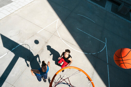 Two young men play basketball on an outdoor court. One looks up at the hoop as a basketball descends towards it. Strong shadows fall across the concrete.