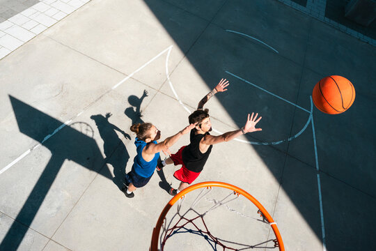 Two young athletes compete in a basketball game on an outdoor court. One jumps to shoot the ball, while the other defends, attempting to block the shot near the hoop.