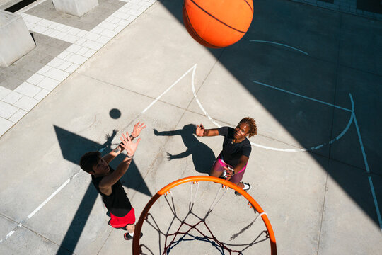 Two diverse friends play basketball on an outdoor court. A man reaches for the ball as it flies towards the hoop, while a woman watches the action.