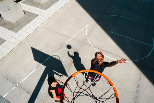 Two young people enjoy a game of basketball on an outdoor court. One player jumps to shoot or block the ball, while the other watches from below the hoop on a sunny day.