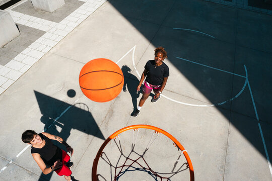 Two diverse women play basketball on an outdoor court. A basketball is in mid-air, heading towards the hoop. Both women look up, enjoying their game under the sun.