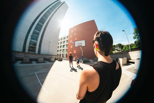 Young people play basketball on an urban court under a bright sun. One person watches the game from the foreground. Modern buildings surround the outdoor court.