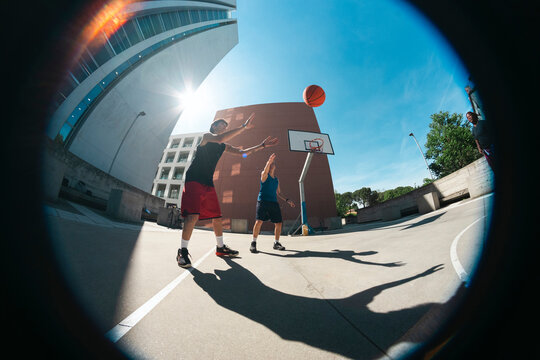 Two young men are actively playing basketball on an outdoor court under a bright sky. One player shoots the ball towards the hoop as the other attempts to block the shot.