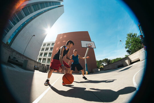Two young women play basketball on an outdoor court under a bright sky. One dribbles the ball while the other defends. Modern buildings surround the urban setting.