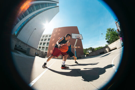 Two young men play basketball on an outdoor court. One dribbles the ball, moving past his defender. Bright sunlight casts long shadows on the concrete, surrounded by modern architecture.