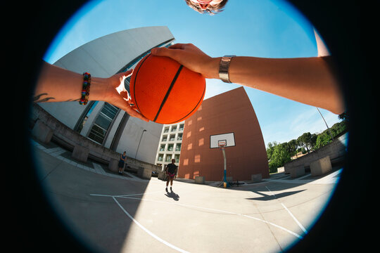 A first-person view of hands holding a basketball on an outdoor court. Two people stand in the background. Modern architecture and a clear sky complete the urban scene.