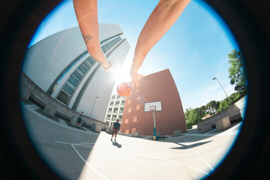 A person holds a basketball, captured from a low fisheye perspective on an outdoor court. Modern buildings and a hoop stand under a bright sky. Two other individuals are nearby.