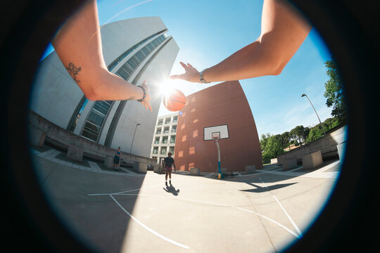 A person's arms are extended, preparing to shoot a basketball on an outdoor court. Another player stands nearby under a bright sky with modern buildings.