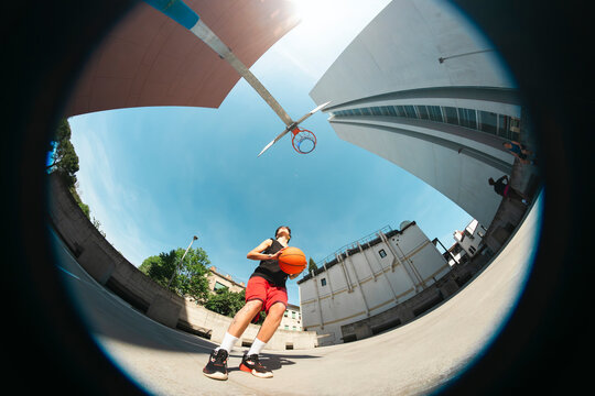 A young athlete in a black tank top and red shorts holds a basketball on an outdoor court. The fisheye perspective captures the hoop, urban buildings, and a bright sunny sky.