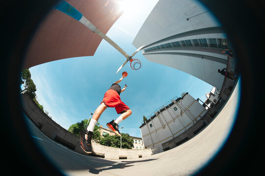 A male athlete in red shorts leaps towards a basketball hoop, attempting a dunk on an outdoor court. The wide-angle view emphasizes the dynamic motion against a clear blue sky and urban architecture.