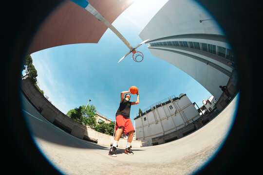 A young male athlete in a black shirt and red shorts holds a basketball, preparing to shoot on an outdoor court. The fisheye perspective captures the urban setting under a bright sky.