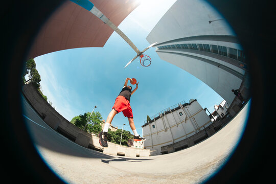 A young athlete in a black tank top and red shorts jumps high to dunk a basketball on an outdoor court. The clear blue sky and modern buildings provide a vibrant backdrop for the dynamic action.