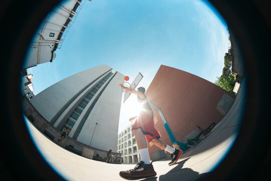 Fisheye view of a man playing basketball outdoors, shooting the ball towards the hoop. Urban setting with modern architecture under a clear sky.