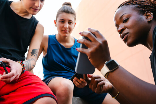 Three young women, two Caucasian and one Black, are outdoors, focused on a smartphone. They are sharing a moment, viewing content together on the mobile device.