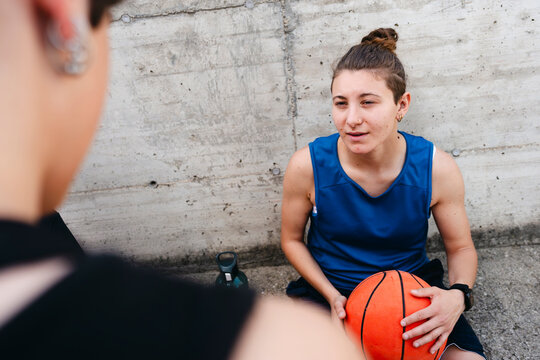Young woman sits on ground, holding a basketball. She wears a blue tank top and shorts, conversing with a friend. Taking a break from playing outdoors against a concrete wall.