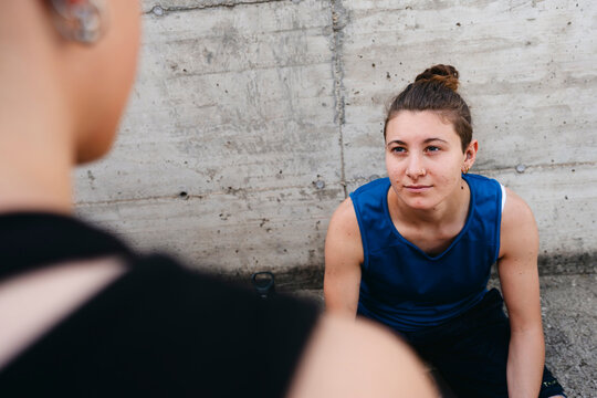 A young woman in athletic attire sits against a concrete wall, looking at another person. Her hair is in a bun, and she wears a nose ring. This shows a moment of pause or conversation.