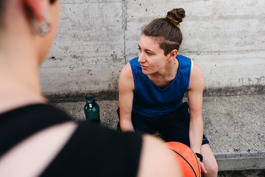 A young person with a bun hairstyle sits on a concrete surface, holding a basketball. They are looking at another person in the foreground, taking a break from a game. A water bottle is beside them.
