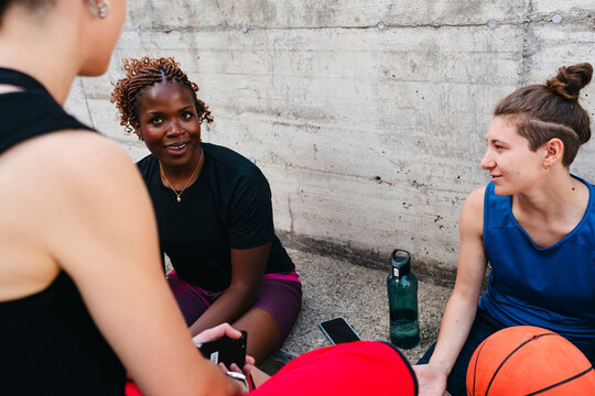 Three diverse women athletes rest against a concrete wall after a game. They are chatting and looking at a phone, with a basketball and water bottle nearby.