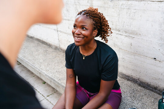 A smiling young Black woman with braided hair sits on a concrete bench, looking at someone out of frame. She wears athletic shorts with a basketball logo, taking a break outdoors.