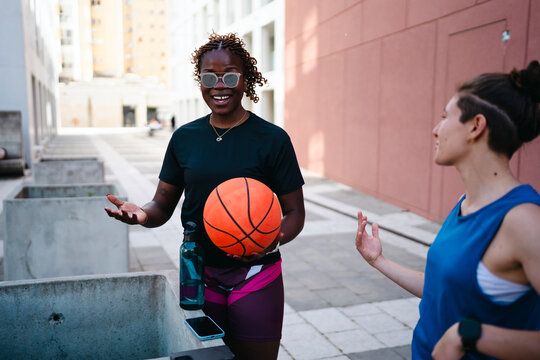 Two young women, one holding a basketball, engage in a lively outdoor conversation. The smiling woman gestures while the other listens, both enjoying their time together in an urban setting.